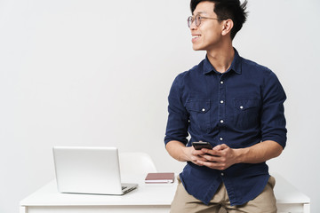 Photo of successful asian businessman sitting at table and holding smartphone while working with laptop in office