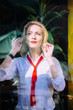 © Iryna - Vertical portrait of blond stylish woman in white shirt with red lips and tie trough the window with fresh green plants on background. Lifestyle portrait concept.