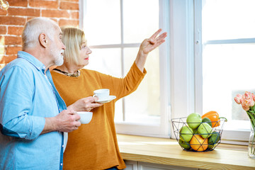 Wall Mural - Portrait of a lovely senior couple standing together with coffee cups, looking out the window at home