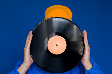 young man in a blue sport 90s style jacket and yellow hat holding vinyl record, man hides his face under vinyl disc