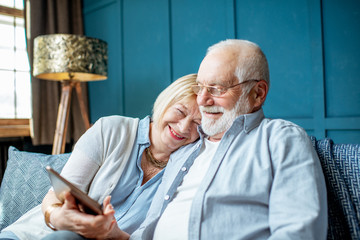 Wall Mural - Lovely senior couple dressed casually using digital tablet while sitting together on the comfortable couch at home