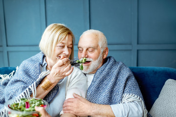 Wall Mural - Lovely senior couple eating healthy salad while sitting wrapped with plaid on the couch at home