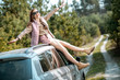 © rh2010 - Young woman enjoying the trip while sitting on the car roof on a picturesque road in the woods