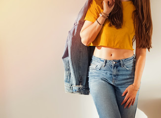 Young woman wearing yellow t-shirt and jeans and holding denim jacket on her shoulder near white wall.