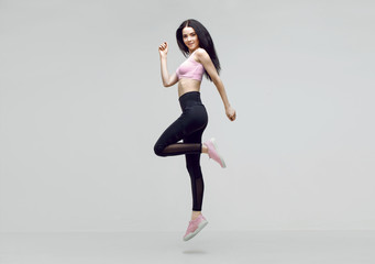 Amazing woman in trendy sportswear jumping. Smiling beautiful slim brunette young girl in fashion leggings and pink top expressing happy emotions. Gray background. Sport, fitness, lifestyle concept.