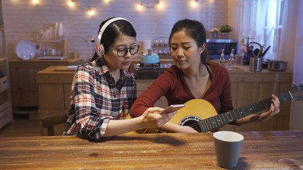 two young asian girls playing music together in house kitchen at night. woman with headphones showing friend cellphone with song app looking lyrics while roommate playing guitar. group enjoy female