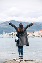 A Trendy Girl Stands Off The Coast Of The Sea And Looks Into The Distance And Raises Her Hands Up Showing How Happy She Is.