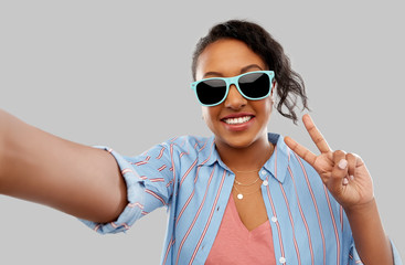 Wall Mural - people, summer and gesture concept - happy african american young woman in sunglasses taking selfie and showing peace hand sign over grey background