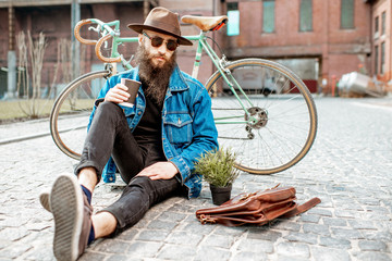 Wall Mural - Portrait of a bearded hipster dressed stylishly with hat and jacket sitting with retro bicycle on the street on the urban background