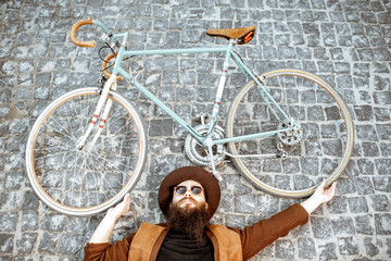 Wall Mural - Portrait of a stylish bearded man in hat lying with retro bicycle on the pavement street, view from above