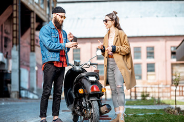 Wall Mural - Stylish young man and woman talking together, standing near the retro moped outdoors on the industrial urban background