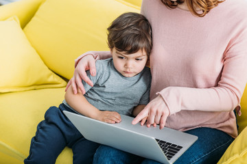 Wall Mural - cropped view of woman with adorable son using laptop while sitting on yellow sofa at home