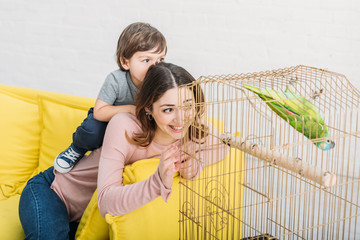 Wall Mural - smiling mother and son looking at green parrot in bird cage at home