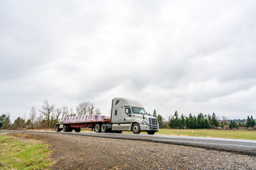 Big rig semi truck transporting fastened commercial cargo on flat bed semi trailer going on the road in overcast weather