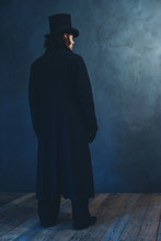 Edwardian Man In Long Black Coat And Hat Standing Towards Grey Wall.