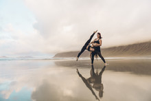 Side View Of Two Barefoot Females In Sportswear Performing Exercise On Wet Sand Near Sea On Overcast Day