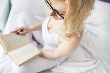 Wall Mural - Attractive blonde woman in glasses with black rim sits on the edge of the bed at home with a book and reads