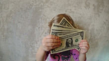 A Small Child Holds A Pack Of One Dollar Banknotes With Two Hands Fanning Out. Background Old Concrete Wall. 