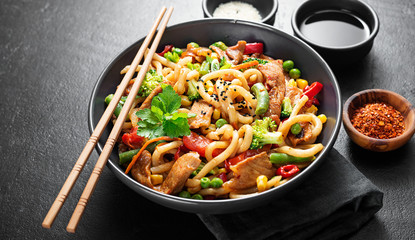 Wall Mural - Udon stir fry noodles with pork meat and vegetables in a dark plate on black stone background.