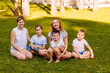 © Alyona - happy women and children sit on the green grass and look at the camera. Big happy family, two mothers and three children in white t-shirts and shorts.