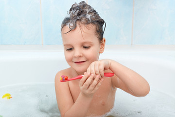 Wall Mural - Shot of little child brushing her teeth while taking bath, charming wet lady holds red tooth brush, has foam on her hair, having fun while washing, glad to clean teeth by herself, water procedures.