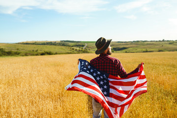 4th of July. Fourth of July. American with the national flag. American Flag. Independence Day. Patriotic holiday. The man is wearing a hat, a backpack, a shirt and jeans. Beautiful sunset light. 