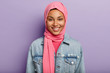Studio shot of glad Eastern female has Islamic religion, covered head with pink veil, smiles gently, shows white teeth, isolated against violet wall expresses positive feelings and emotions. Ethnicity