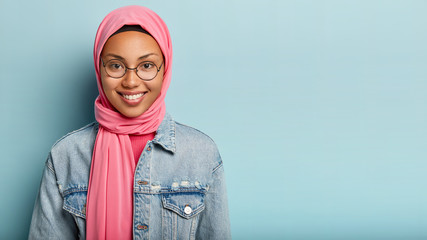 Glad satisfied delighted woman smiles broadly at camera, being in high spirit, looks through optical glasses, has Islamic views, stands against blue studio wall with empty space for your promotion