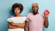 Couple In Love With Dark Skin Awoke Late And Overslept Morning Alarm Clock, Stare At Camera, Hold Pillow, Ringing Bell, Isolated On Blue Background. Early Morning. Harmful Habit To Oversleep