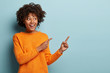 Leinwandbild Motiv Horizontal shot pleased dark skinned female with Afro haircut, points away with both fore fingers, shows blank space for your promotion, isolated over blue background. People, advertisement concept