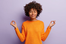 Pleasant Looking Calm Woman Meditates Indoor, Holds Hands In Mudra Gesture, Has Charming Smile, Closed Eyes, Wears Orange Clothes, Models Over Purple Background. Hand Gesture. Meditation Concept