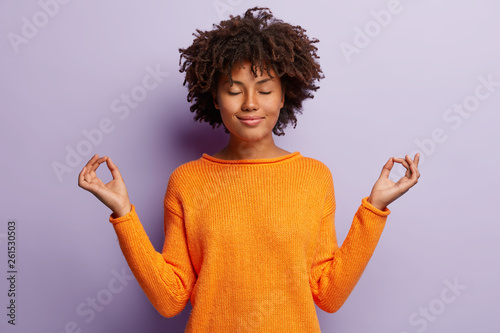 Cuadros en Lienzo Pleasant looking calm woman meditates indoor, holds hands in mudra gesture, has charming smile, closed eyes, wears orange clothes, models over purple background