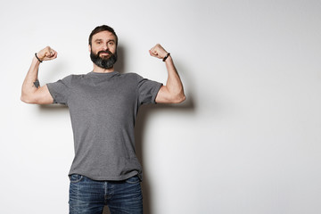 Wall Mural - Positive brutal tattooed bearded guy poses in blue jeans and blank gray t-shirt premium summer cotton, on white background mockup.