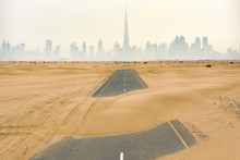 Aerial View Of A Deserted Road Covered By Sand Dunes In The Middle Of The Dubai Desert. Dubai Skyline With The Burj Khalifa Surrounded By Fog In The Background. Dubai, United Arab Emirates.