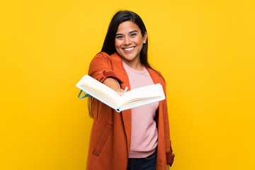 Wall Mural - Young Colombian girl over yellow wall holding a book and giving it to someone