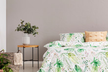 Green Flower In Grey Vase On Wooden Bedside Table Next To Comfortable Bed With Floral Bedding, Copy Space On The Empty Grey Wall