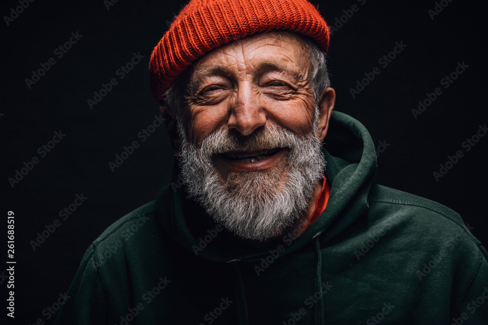 Close up portrait of happy 70-year-old optimist man with smiling wrinkled face, dressed in hipster orange hat and green hoodie, isolated over black background. Positive and cheerful at any age. - obrazy, fototapety, plakaty 