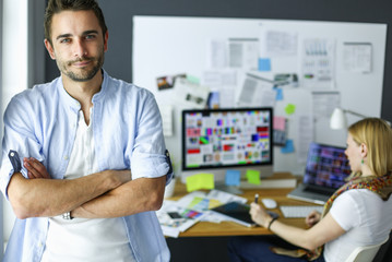 Poster - Portrait of young designer in front of laptop and computer while working. Assistant using her mobile at background.