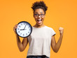 Joyful girl holds clock, raises fist and shouts loudly. Photo of african american girl wears casual outfit on orange background. Emotions and pleasant feelings concept.