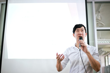 Portrait of young handsome Asian male speaker publicly speaking on stage to group of audience with white board behind