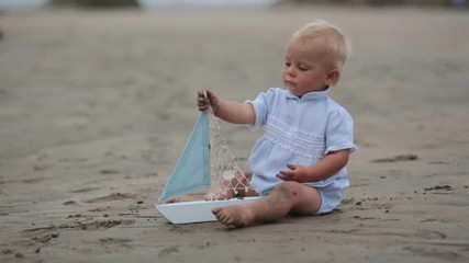 Wall Mural - Baby boy sitting on the beach near the water and plays with a toy ship and teddy bear