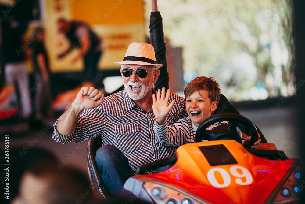 Grandfather and grandson having fun and spending good quality time together in amusement park. They enjoying and smiling while driving bumper car together. - obrazy, fototapety, plakaty 