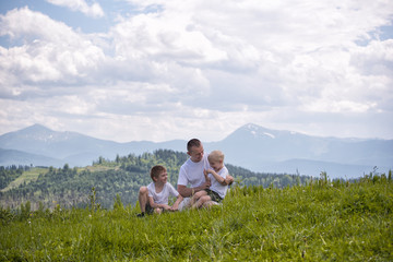 Happy father with his two young sons sitting on the grass on a background of green forest, mountains and sky with clouds. Friendship concept.