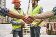 © Friends Stock - We did it! Close up photo of builders shaking hands against cheerful colleagues while working together at construction site. Team work.