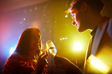 Wall Mural - Smiling beautiful young couple standing in nightclub with disco lights and clinking champagne flutes while celebrating anniversary