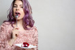 Cute young woman with purple hair eating delicious cheese cake over grey background. Empty space