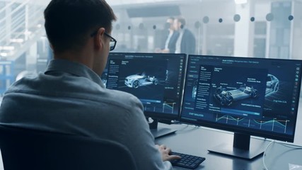 Fototapete - Professional Engineer Works on a Computer with a 3D CAD Software and Tests the Electric Car Chassis Prototype with Wheels, Batteries and Engine Standing in a High Tech Development Laboratory.