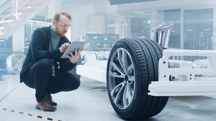 Poster - Engineer with Glasses and Beard Works on a Tablet Computer Next to an Electric Car Chassis Prototype with Wheels, Batteries and Engine in a High Tech Development Laboratory.