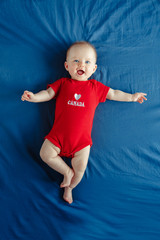 white caucasian smiling baby boy girl with blue eyes lying on bed at home on canada day. newborn inf