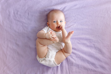 Portrait of cute adorable white Caucasian baby girl boy with blue eyes lying on bed looking at camera and licking sucking fingers fist. View from top above. Happy childhood lifestyle.